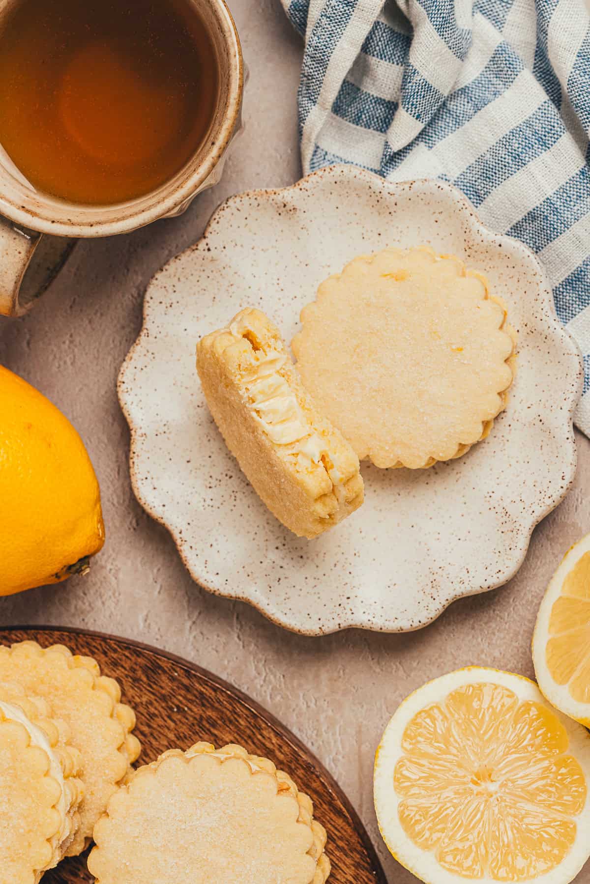 Overhead view of lemon sandwich cookies on a scalloped plate with fresh lemon halves and a cup of tea.