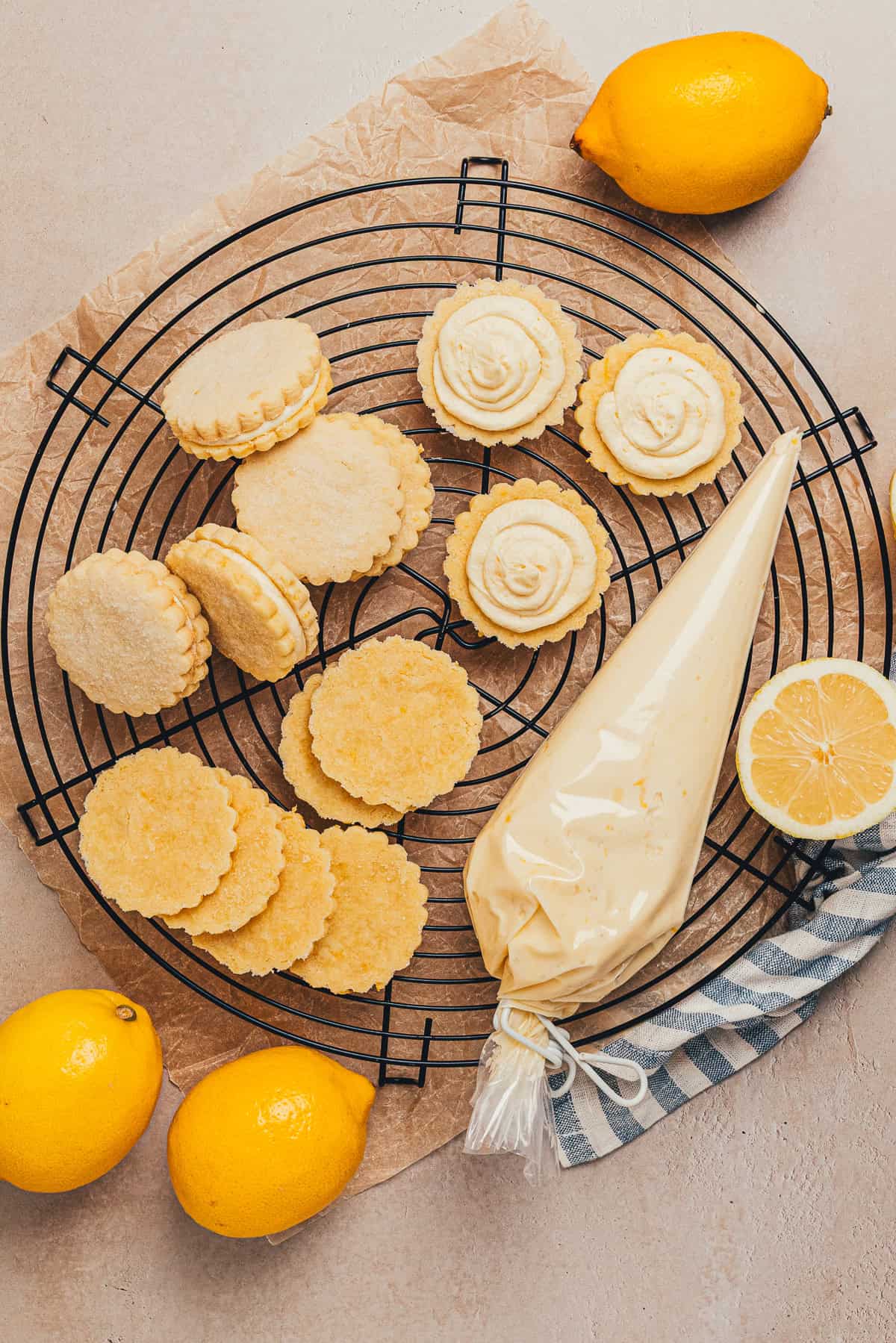 Lemon sandwich cookies being assembled with buttercream, piping bag, and fresh lemons on a cooling rack.