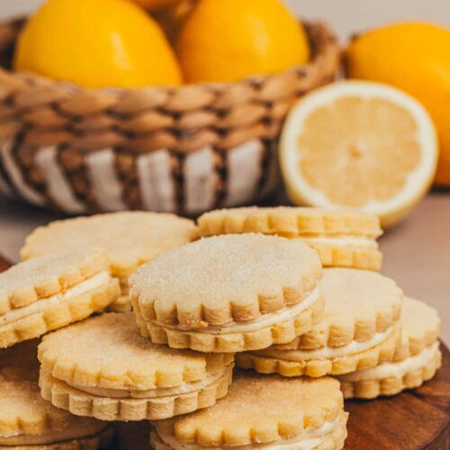Lemon sandwich cookies with buttercream filling on a wooden board with fresh lemons in the background.