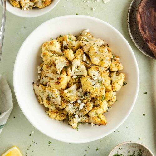 Roasted cauliflower with dill and feta served in a white bowl with lemon wedges and fresh herbs on a light green background.