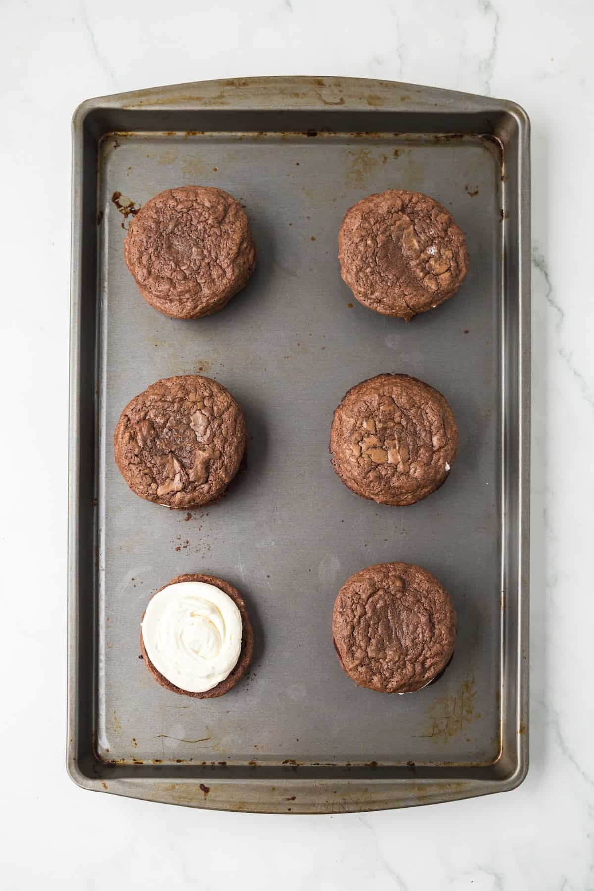 Baked brownie cookies on a baking sheet with one cookie topped with cream filling.