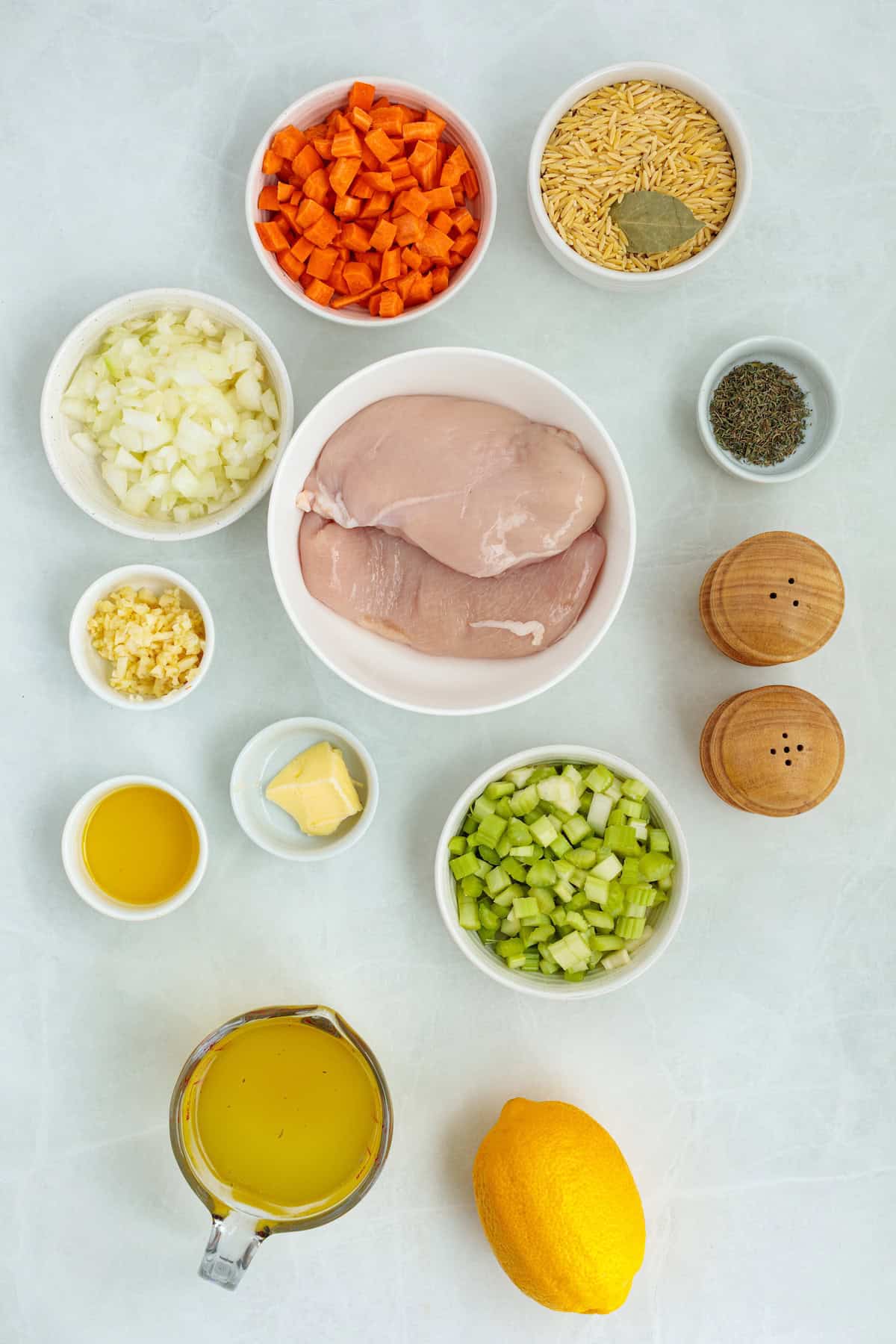 Overhead view of the ingredients for homemade lemon chicken orzo soup, including chicken breasts, carrots, celery, onion, garlic, orzo pasta, chicken broth, butter, olive oil, thyme, lemon, salt, and pepper.