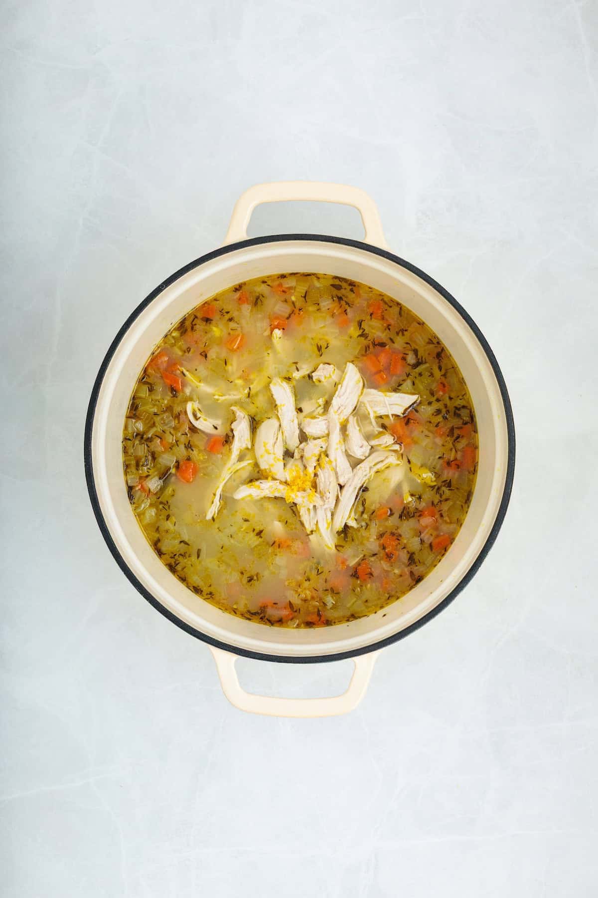 Overhead view of shredded chicken and fresh lemon zest added to seasoned broth with carrots, celery, onions, and herbs before stirring into homemade lemon chicken orzo soup.
