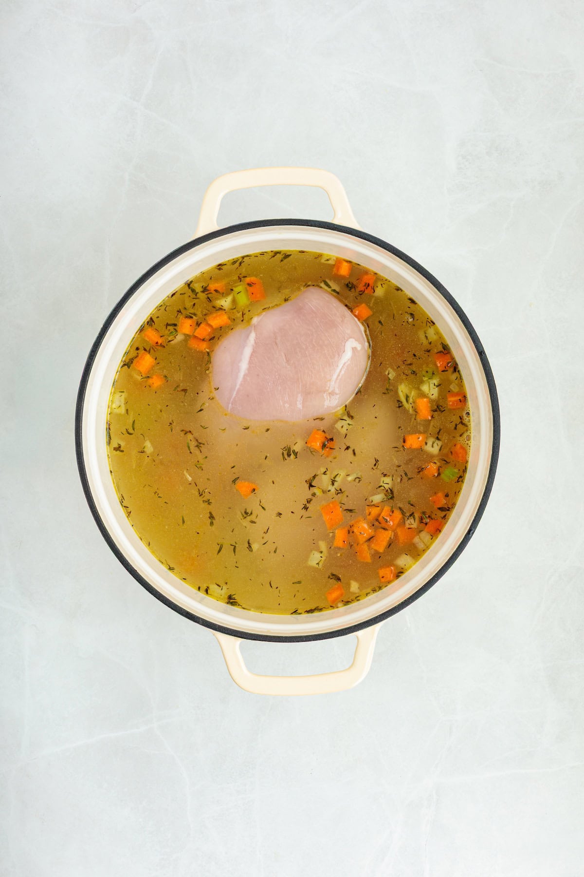 Overhead view of a raw chicken breast simmering in seasoned broth with diced carrots, celery, onions, and herbs in a Dutch oven for homemade lemon chicken orzo soup.