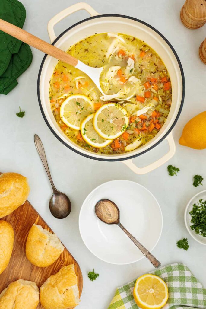 Overhead view of homemade lemon chicken orzo soup ready to serve in a Dutch oven with a ladle, fresh lemon slices, shredded chicken, crusty bread, and serving bowls.