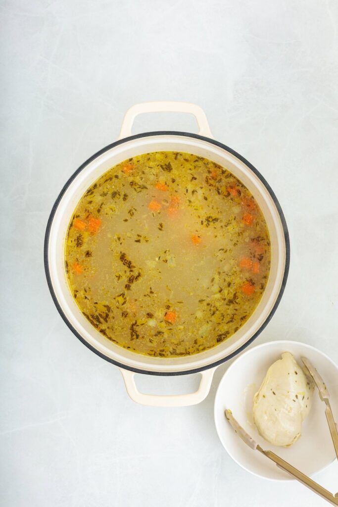 Homemade lemon chicken orzo soup broth simmering with vegetables and herbs in a Dutch oven, with cooked chicken breast set aside before shredding and adding back to the soup.