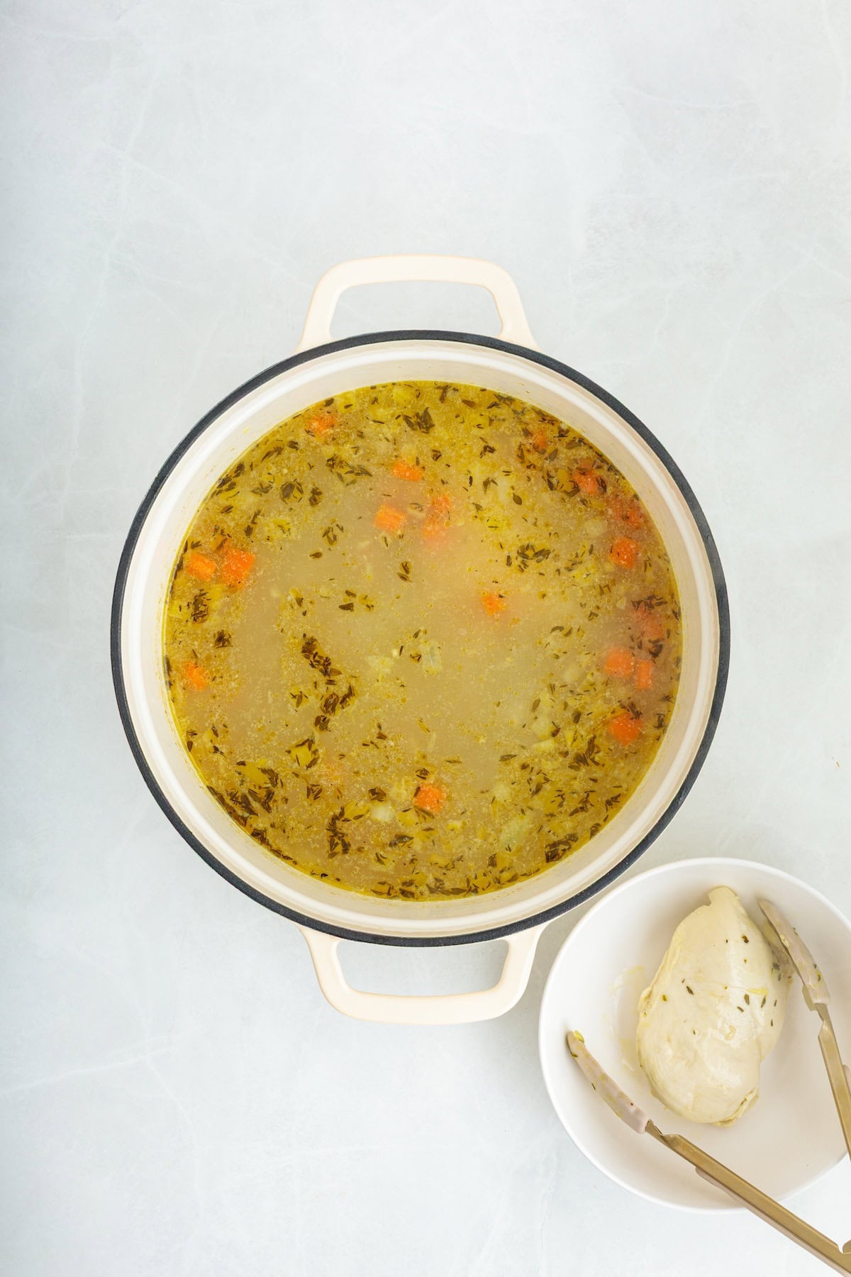 Homemade lemon chicken orzo soup broth simmering with vegetables and herbs in a Dutch oven, with cooked chicken breast set aside before shredding and adding back to the soup.