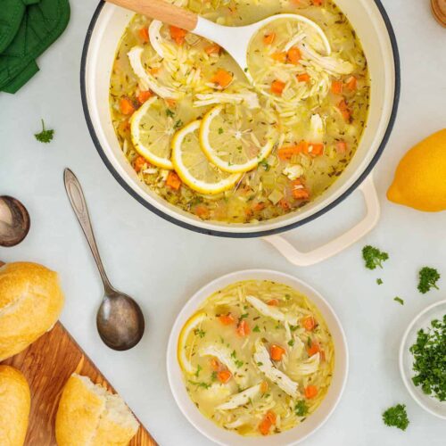 Overhead view of homemade lemon chicken orzo soup in a Dutch oven with a ladle, served with a bowl of soup, crusty bread, fresh parsley, and lemon slices.
