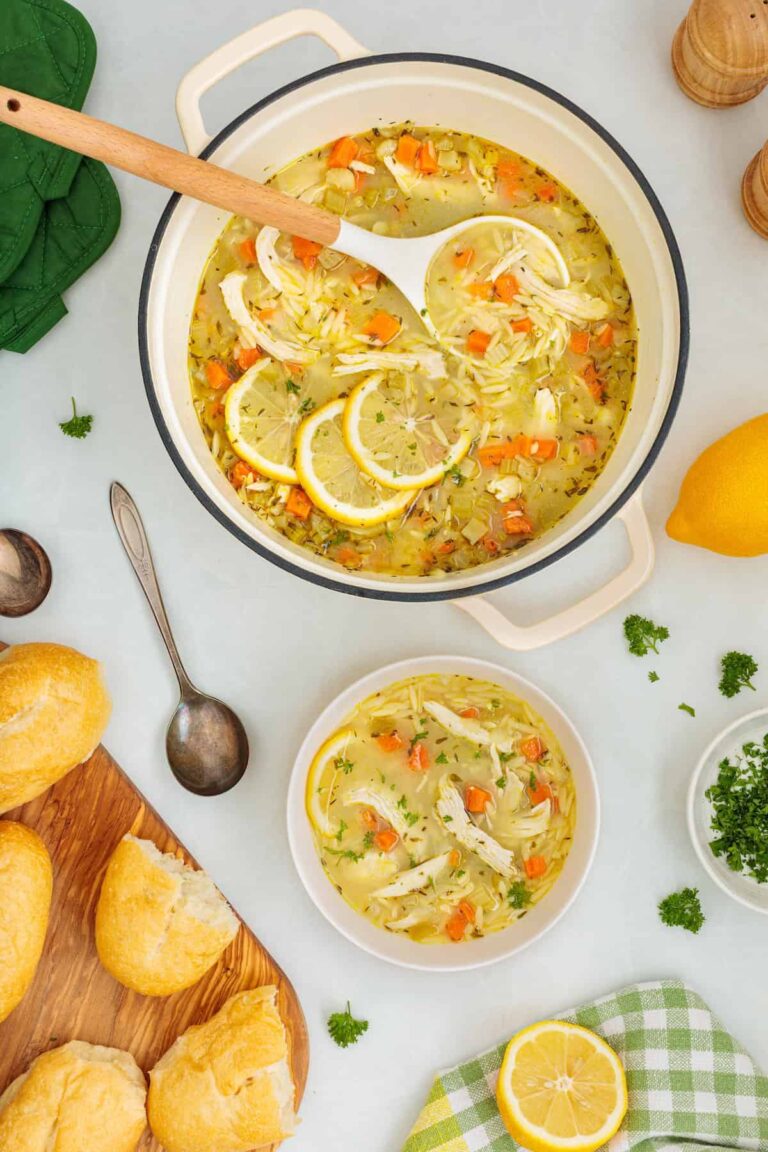 Overhead view of homemade lemon chicken orzo soup in a Dutch oven with a ladle, served with a bowl of soup, crusty bread, fresh parsley, and lemon slices.