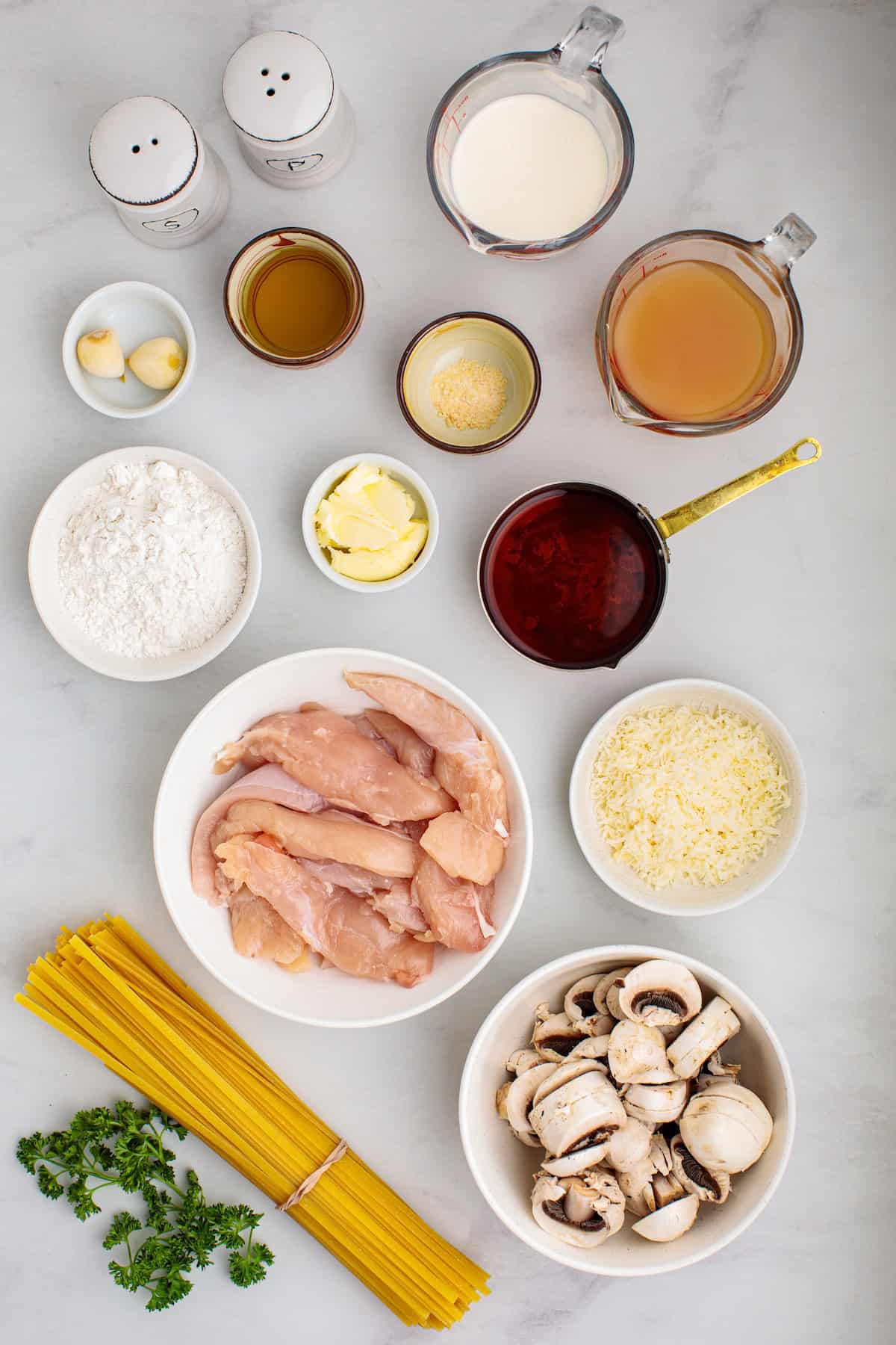 Ingredients for creamy chicken Marsala fettuccine including chicken tenders, mushrooms, pasta, Marsala wine, Parmesan cheese, cream, garlic, and fresh parsley arranged on a marble countertop.